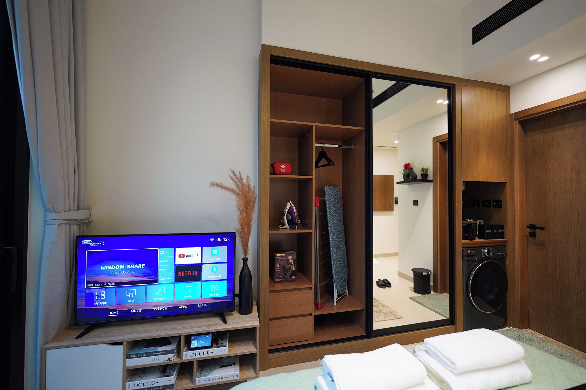 Une chambre d’hôtel moderne équipée d’un téléviseur affichant un écran d’accueil, d’une armoire en bois, d’un lave-linge et de serviettes soigneusement pliées.