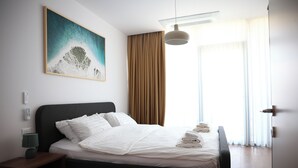 Una habitación de hotel moderna con una cama bien hecha, un cuadro en la pared, una mesita de noche con una lámpara y un ventanal con cortinas.