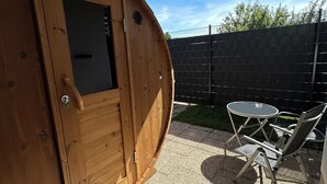 Eine Holzhütte mit Tür und kleinem Fenster, einem runden Tisch mit Stühlen sowie einer überdachten Terrasse.