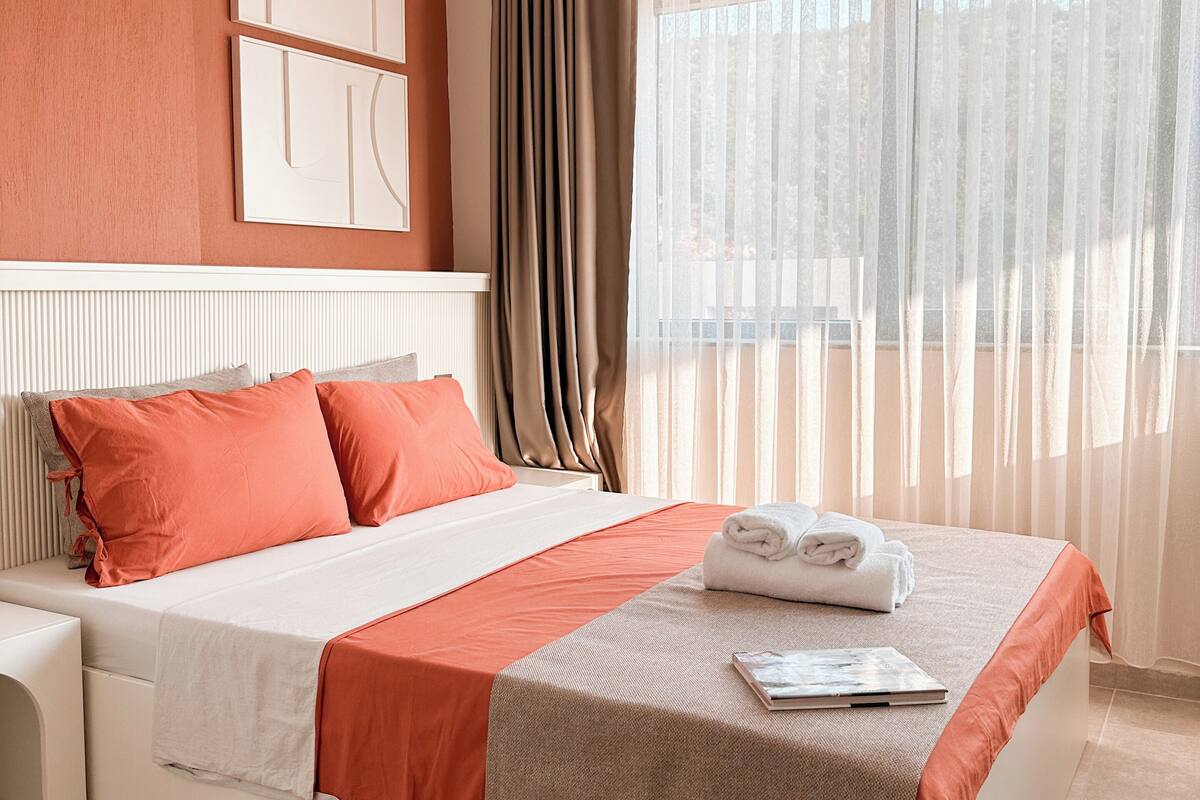 Ein Hotelzimmer mit einem großen Bett, einem Nachttisch, einem Fenster mit leichtem Vorhang und einer Deckenleuchte.