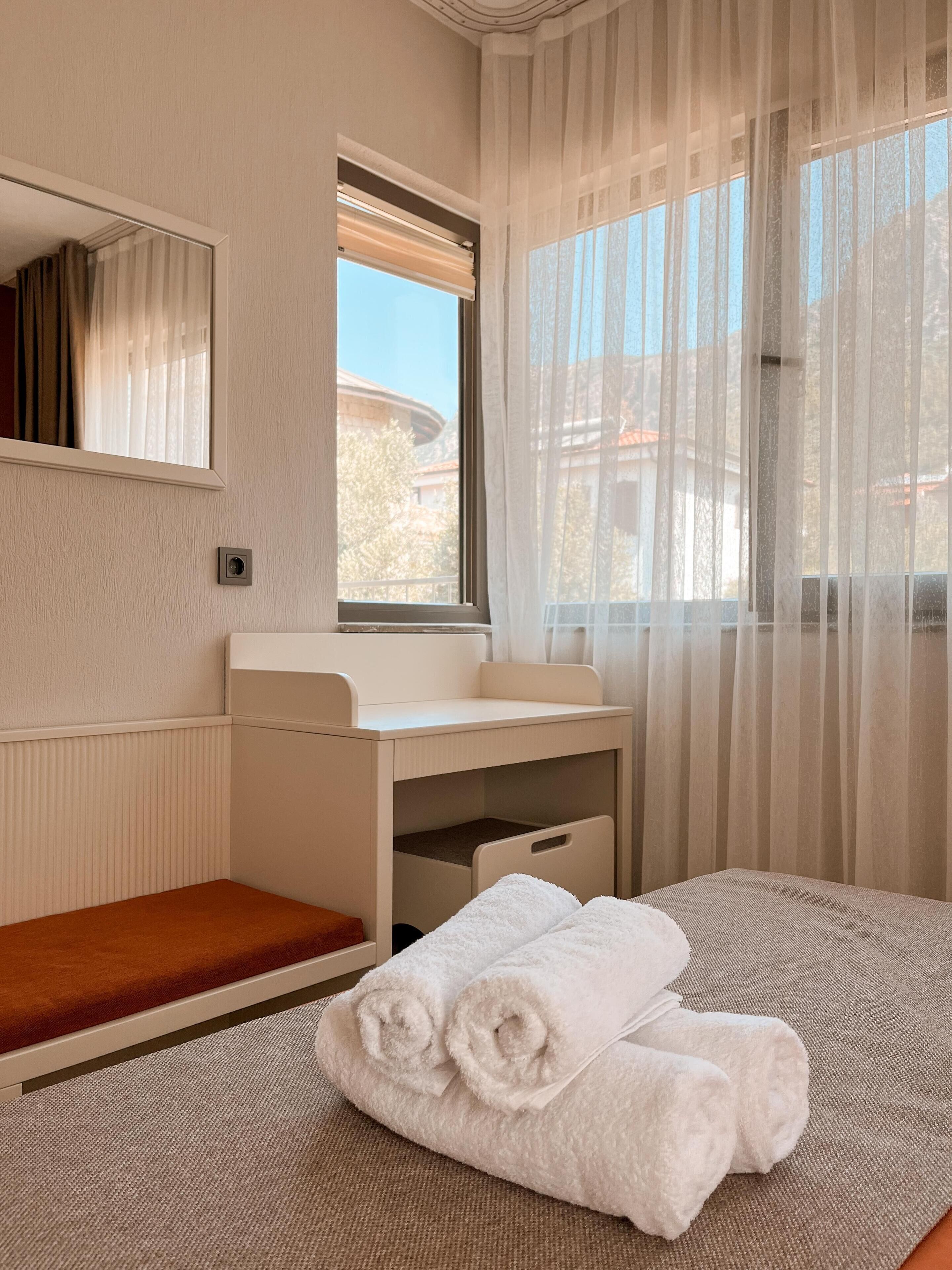 A hotel room with a bed, a small desk with a mirror, a window with sheer curtains, and a view of buildings outside.