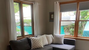 Un salon moderne avec un canapé d’angle gris, des coussins blancs et une grande fenêtre agrémentée de rideaux blancs.