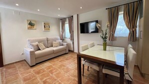 Un salon comprenant un canapé, une table à manger avec des chaises, un téléviseur à écran plat fixé au mur et une fenêtre avec des rideaux.
