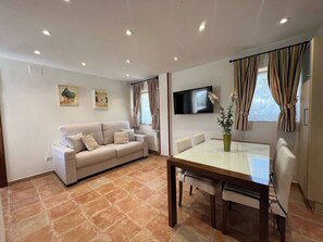 Un salon comprenant un canapé, une table à manger avec des chaises, un téléviseur à écran plat fixé au mur et une fenêtre avec des rideaux.