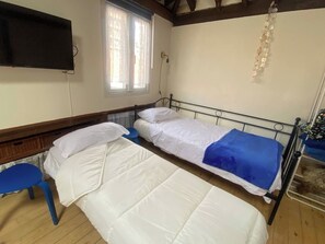Una habitación con dos camas individuales, una silla azul, un televisor montado en la pared y una ventana con cortinas.