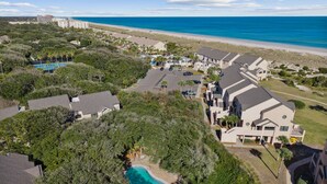 An aerial view of a coastal residential area with a swimming pool surrounded by lush greenery, a parking lot, and houses with a view of the beach and ocean.