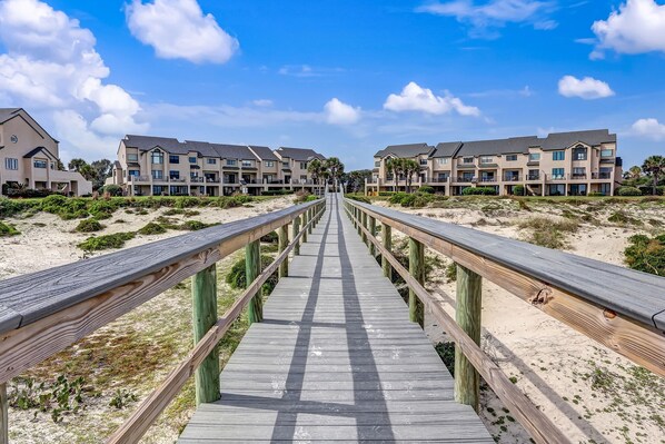 A wooden boardwalk leading to a beachfront building complex with multiple balconies and a clear blue sky.