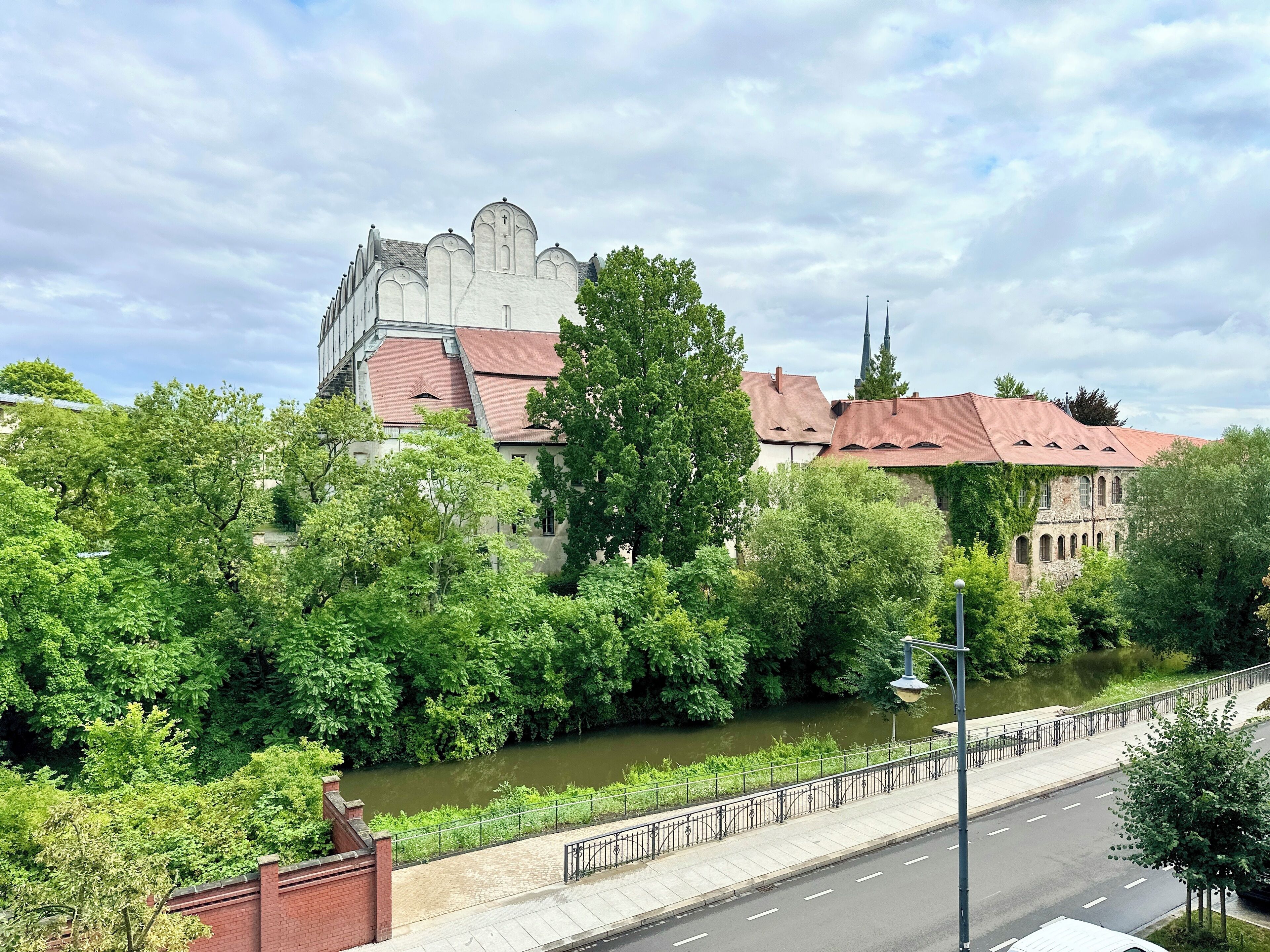 A historic building with a red roof and white walls, surrounded by lush greenery and a river.