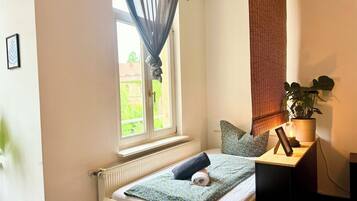 A small, cozy bedroom with a window, a radiator, a bedside table with a plant, and a neatly made bed.