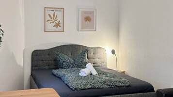 Un lit bien fait, recouvert d’une couverture foncée, avec une tête de lit grise et un oreiller. Deux tableaux encadrés sont accrochés au mur, au-dessus du lit.