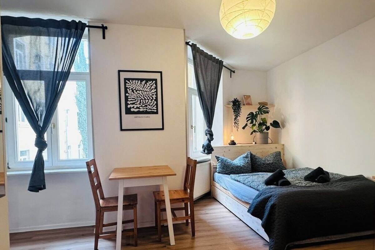Una camera compatta con un letto, una scrivania e una sedia. Si vede una finestra con tende, un'opera d'arte appesa al muro e una pianta sul comodino.