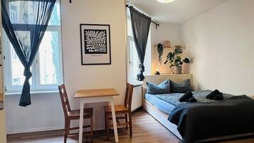 A compact room with a bed, a desk, and a chair. There is a window with curtains, a wall-mounted artwork, and a bedside plant.
