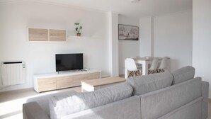 Uma sala de estar moderna com um sofá cinzento, uma TV de tela plana, uma mesa de centro de madeira e um espaço de jantar com uma mesa e cadeiras brancas.