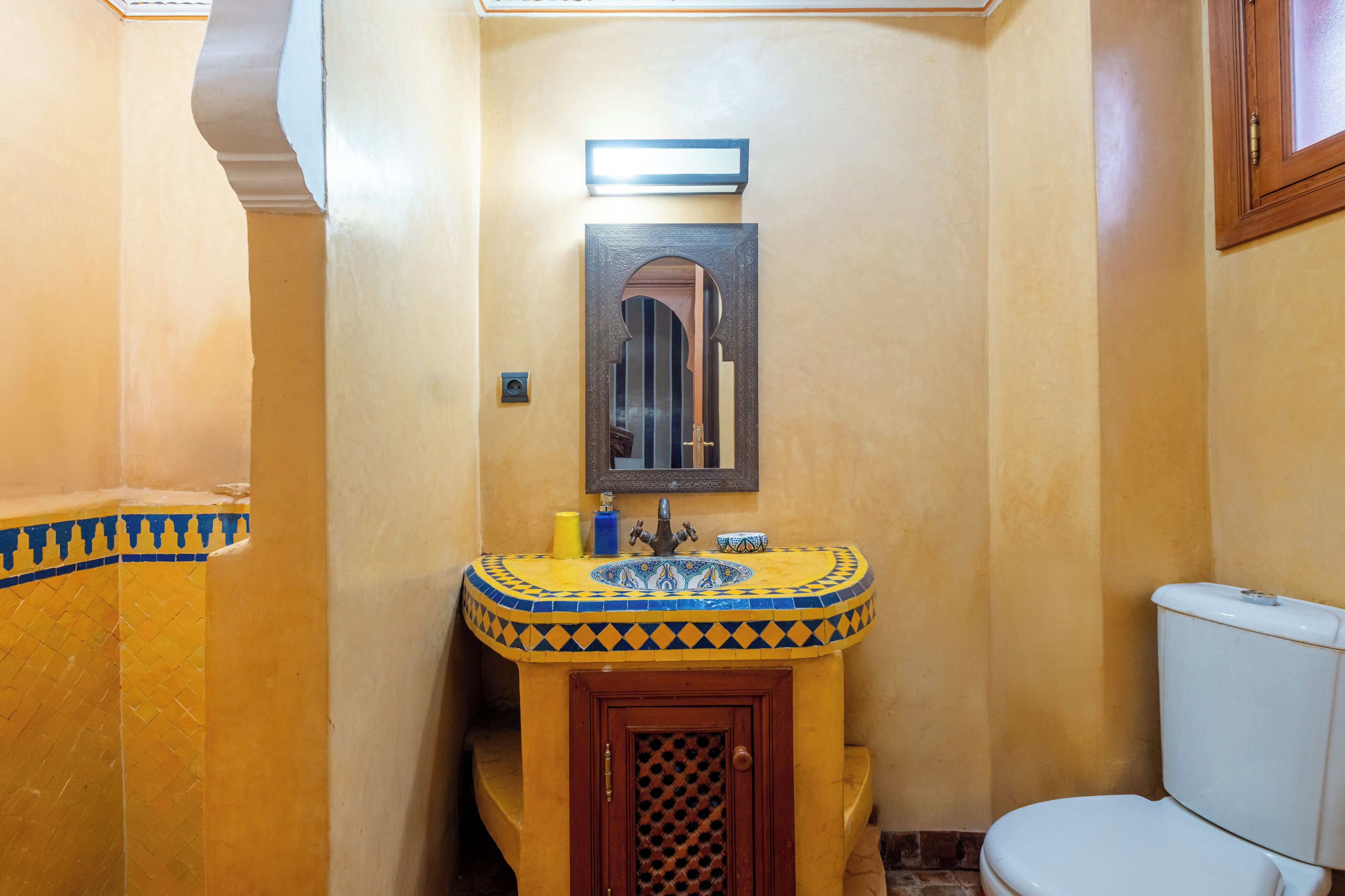 A bathroom with a yellow tiled wall, a sink with a decorative tile pattern, a mirror, and a white toilet.