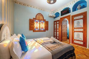 A bedroom with a bed, pillows, a window with stained glass, an air conditioning unit, a television, and a door with decorative panels.