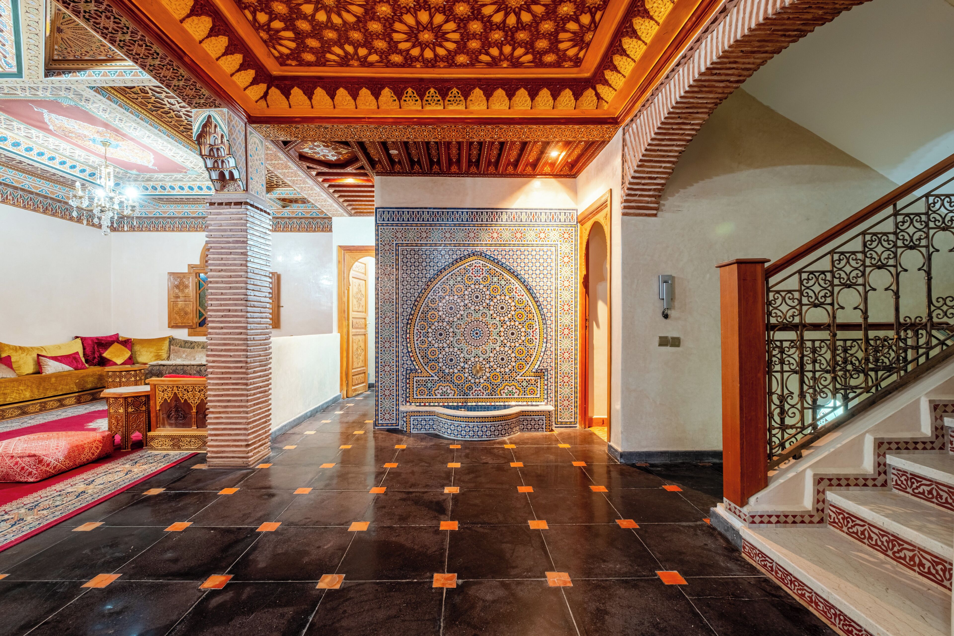 Un vaste hall d’entrée orné de motifs complexes dans les carreaux, un escalier avec des rampes richement décorées et un coin salon confortable agrémenté de coussins.