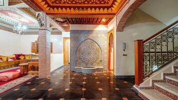 Un vaste hall d’entrée orné de motifs complexes dans les carreaux, un escalier avec des rampes richement décorées et un coin salon confortable agrémenté de coussins.