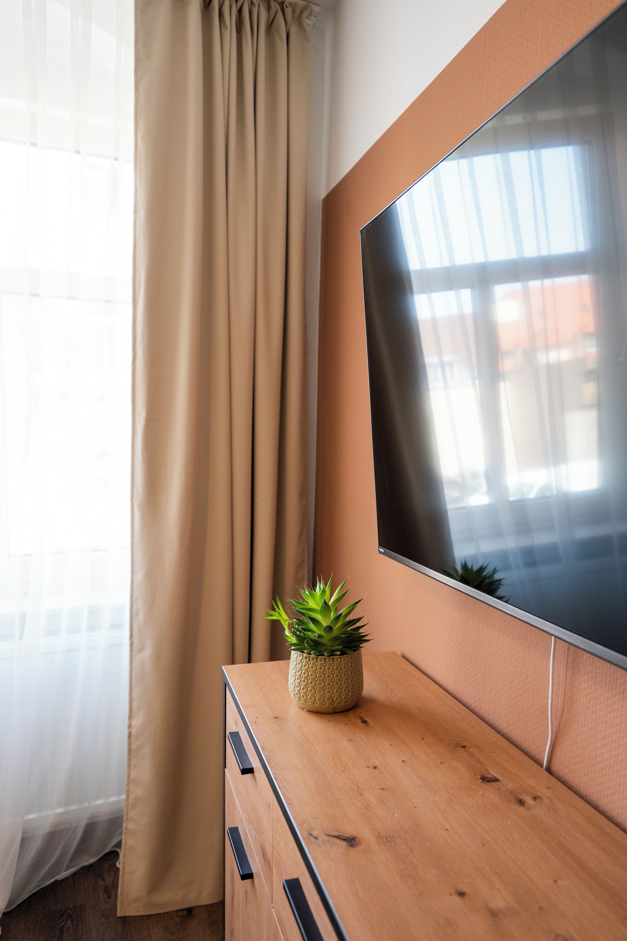 Ein Zimmer mit einem hölzernen Schminktisch, einer Topfpflanze, einem großen Flachbildfernseher und einem Fenster mit Vorhängen.