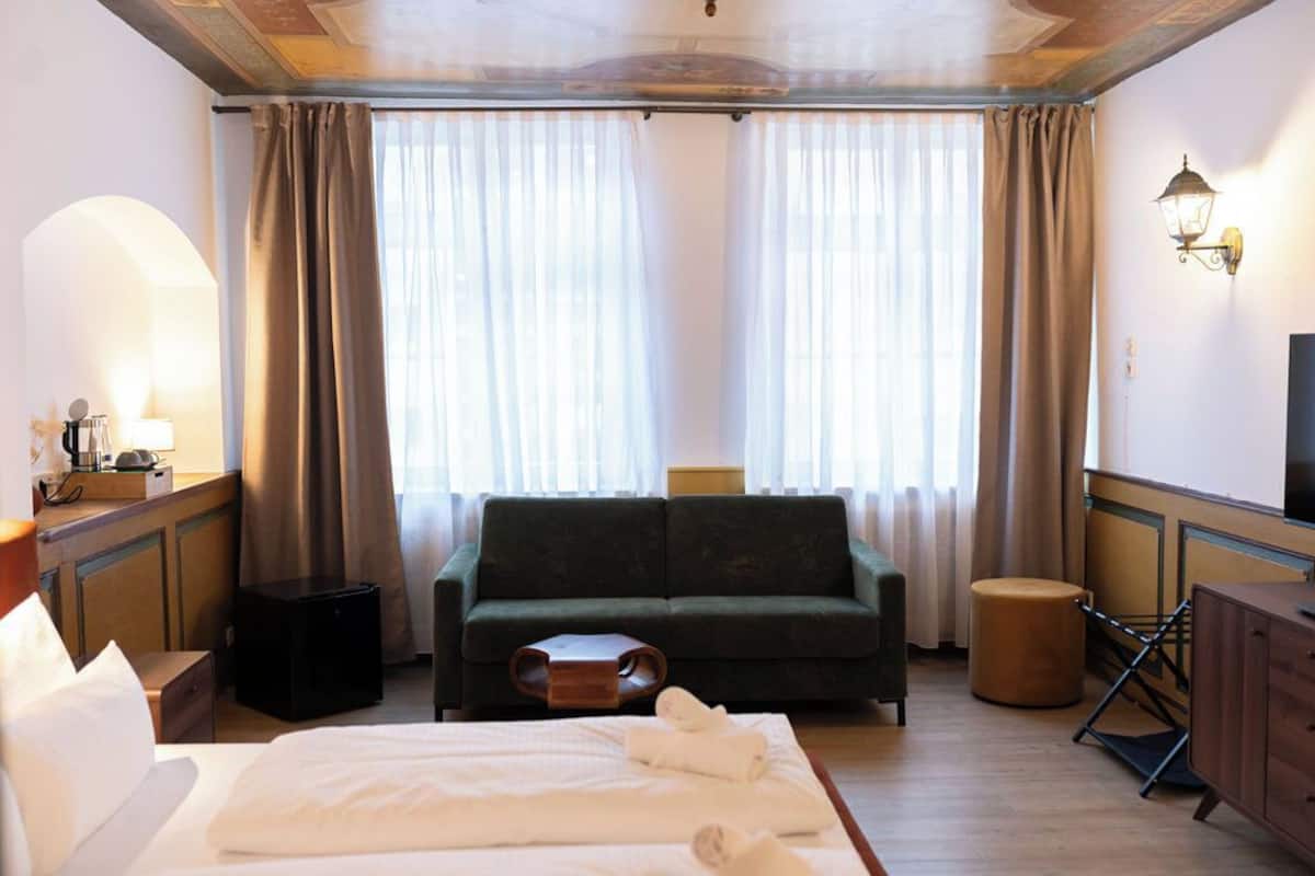 Habitación de hotel con cama, sofá verde, pequeña mesa de madera, televisor y ventana con cortinas.