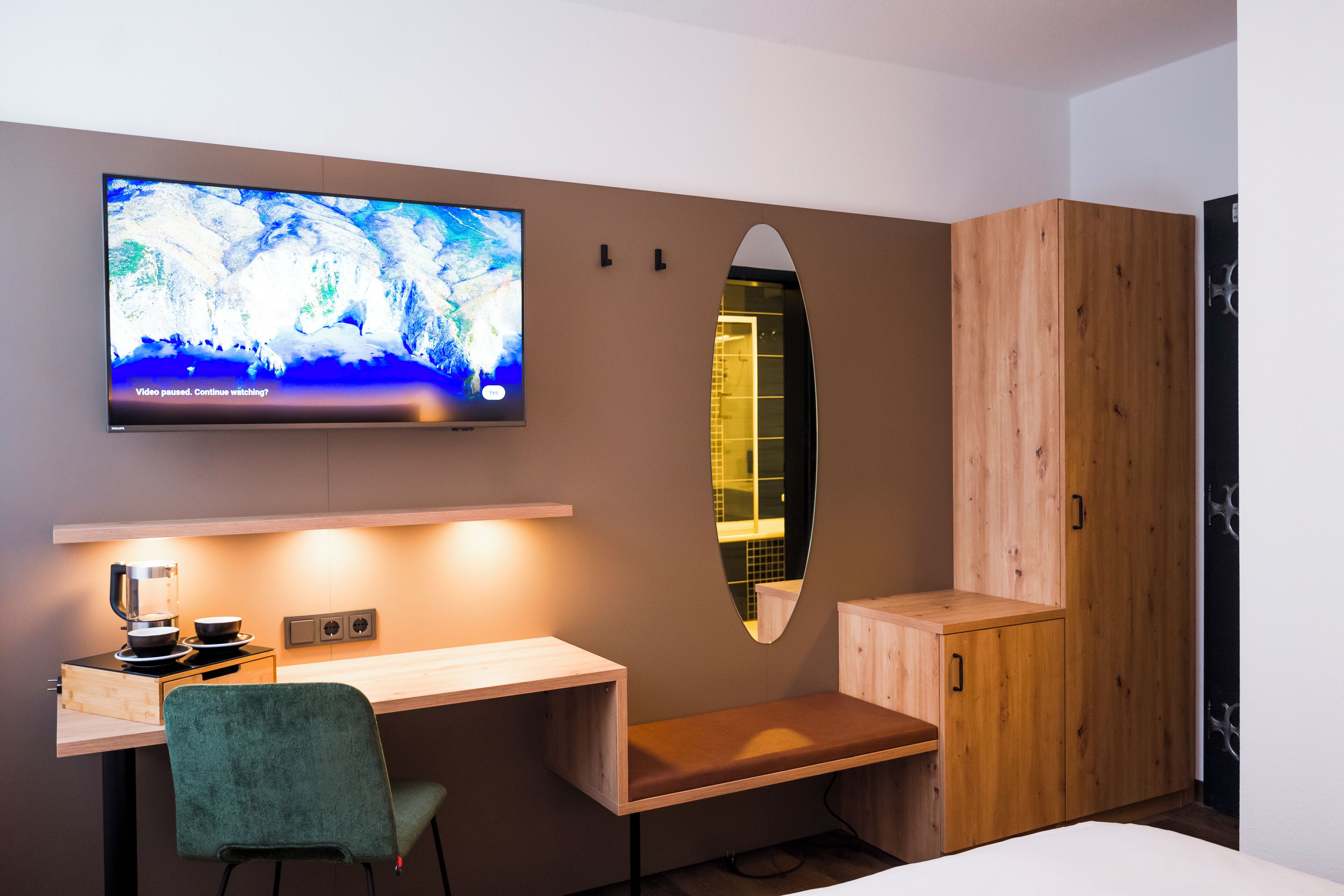 A modern bedroom with a TV mounted on the wall, a wooden desk with a chair, a bench, and a wardrobe.