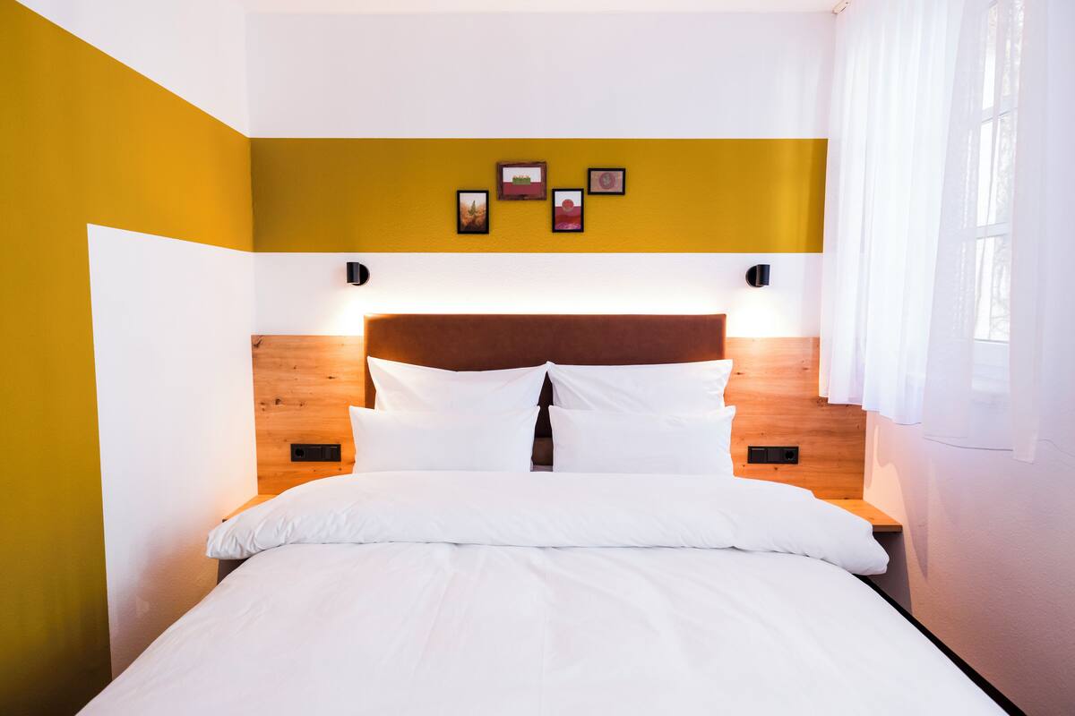 Una cama bien tendida con sábanas blancas en una habitación con paredes amarillas y blancas, un cabecero de madera y un pequeño estante de pared con marcos de fotos.