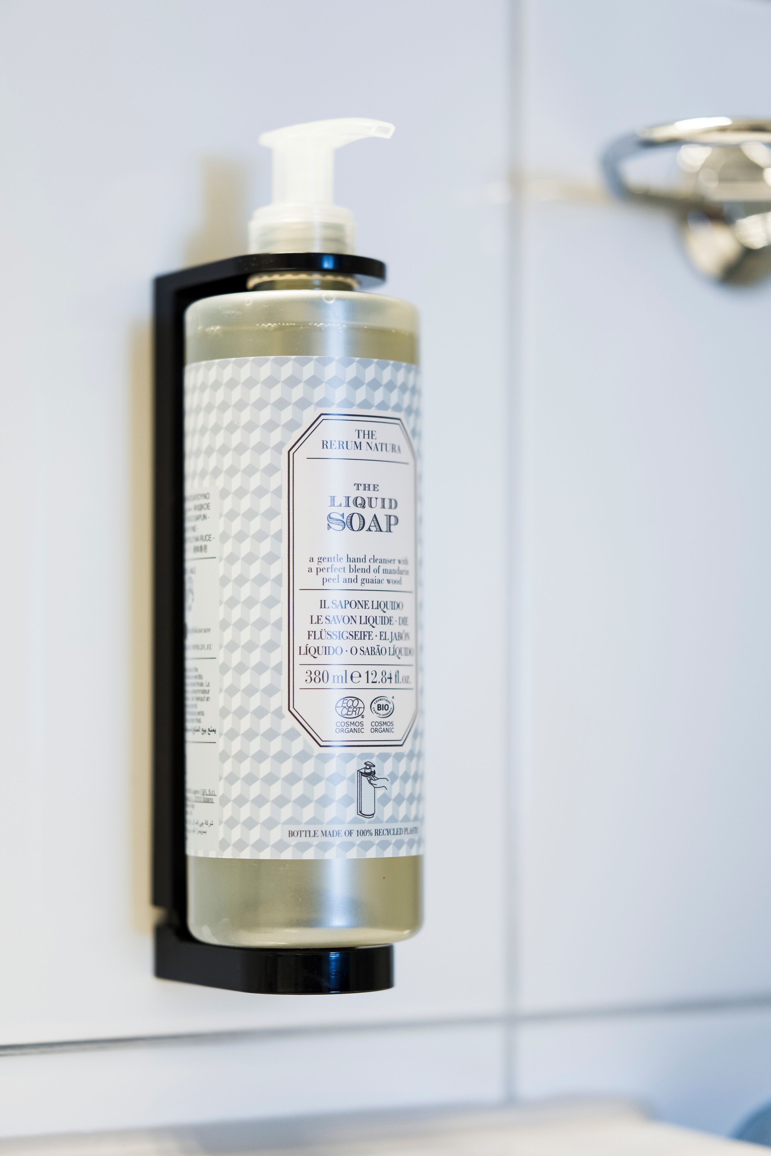 A bottle of liquid soap with a pump dispenser, labeled as organic and made from recycled materials, mounted on a white wall.