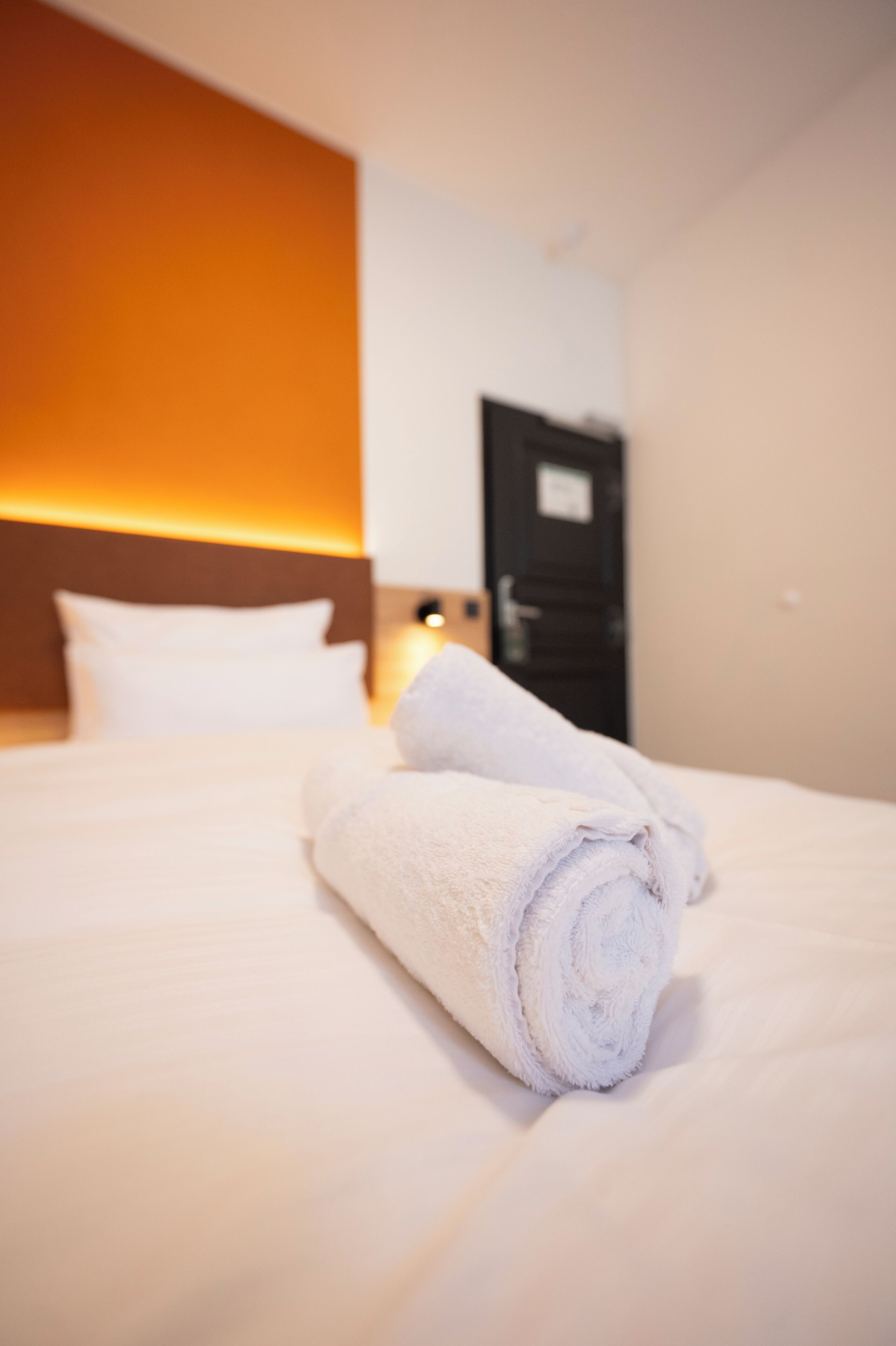 A neatly made bed with rolled white towels, a white headboard, and a door in the background.