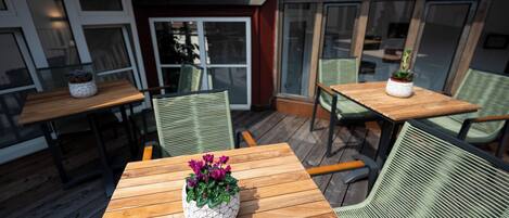 An outdoor patio area with wooden tables and chairs, potted plants, and a view of the building's interior through large windows.