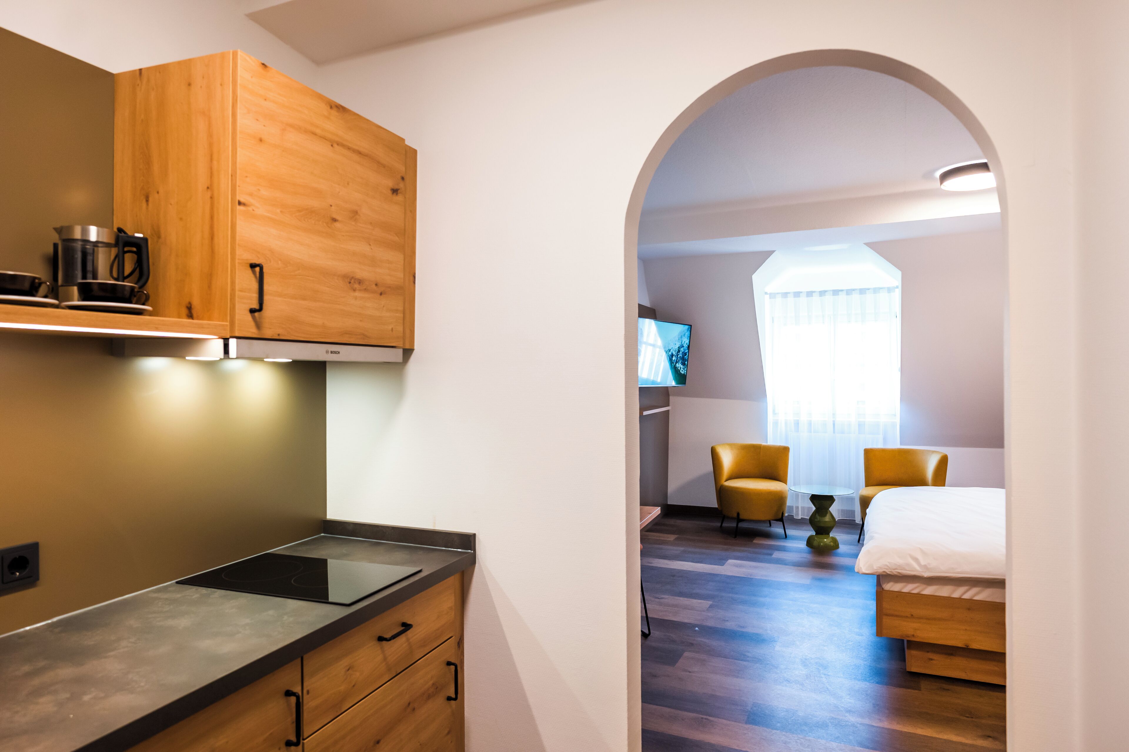 A modern kitchen with wooden cabinets and a built-in stove, leading to a bedroom with a bed, two chairs, and a TV.