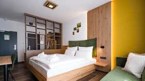 Une chambre d’hôtel moderne avec un lit en bois, un canapé vert, une bibliothèque intégrée en bois et un mur d’accent jaune.