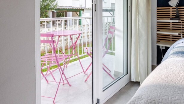 Una habitación con puerta de vidrio corrediza que da a un balcón exterior con sillas rosas y una mesa.