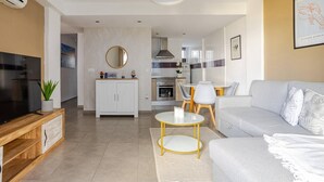 Una sala moderna con sofá, mesa de centro y televisión. Se observa una cocina al fondo con zona de comedor.