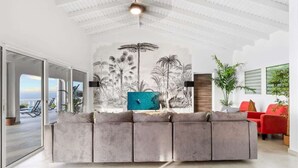 Un salon moderne avec un canapé d’angle gris, un fauteuil rouge, une grande fresque murale et une vue sur la mer grâce à de grandes fenêtres.