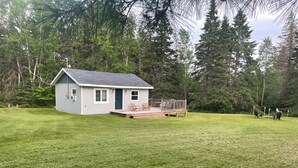 A small, single-story house with a porch and a deck, surrounded by a well-maintained lawn and dense forest.