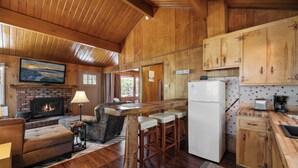 A cozy cabin interior with a fireplace, wooden bar stools, a sofa, and a kitchen area with wooden cabinets and a refrigerator. - Chateau Rouge (Big Bear Lake)
