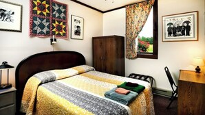 A bedroom with a bed, a wooden wardrobe, a desk with a lamp, and framed pictures on the walls.