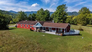 A red building with a porch and outdoor seating, surrounded by a grassy field and trees, with mountains in the background.