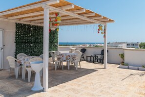 A covered outdoor seating area with a table and chairs, a pergola, and a view of the sea.