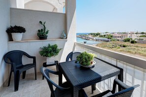 A balcony with a table and chairs, potted plants, and a view of the sea and buildings. - Appartamento Cala Pescatori (Villanova)