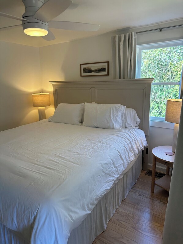 Een slaapkamer met een bed, een plafondventilator, een raam met gordijnen, een bedlampje en een klein tafeltje.