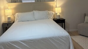 Un llit ben arrenjat amb llençols blancs, dues làmpades laterals, un ventilador de sostre i una finestra amb cortines.