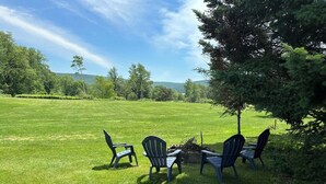 Un espace extérieur verdoyant avec quatre chaises Adirondack bleues disposées autour d’un petit foyer.