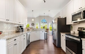 A spacious kitchen with white cabinets, a large island, stainless steel appliances, and a view of greenery through a window. - The Chick-Inn: Monthly Discounts, Groups, Parking! (Southport)