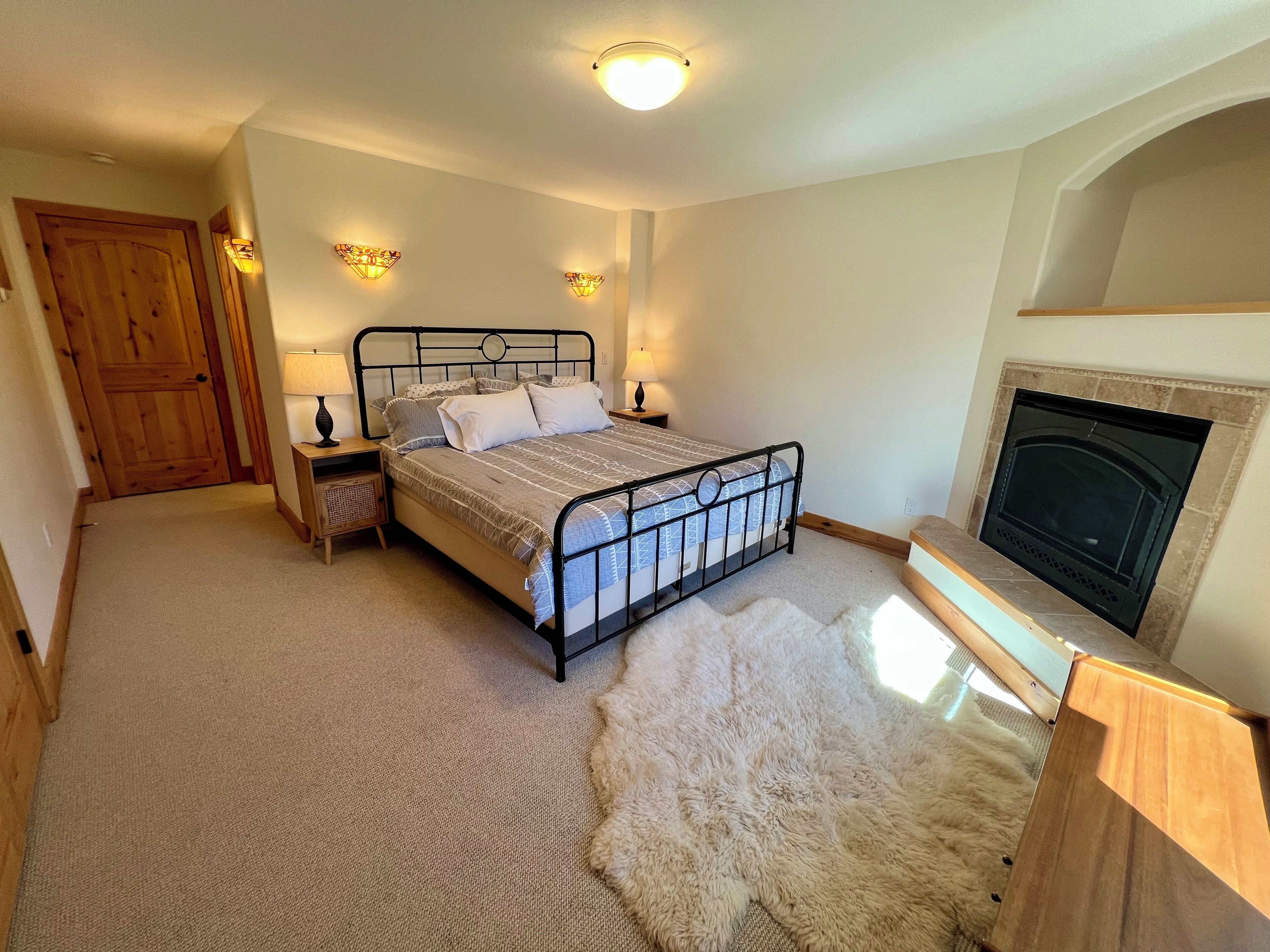 A bedroom with a bed, bedside lamps, a wooden door, a fireplace, and a fur rug.