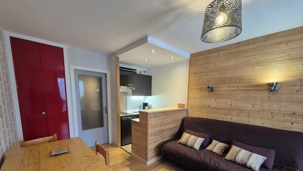 Un espacio habitable compacto con una mesa de comedor de madera, un sofá con cojines, una puerta roja y una zona de cocina con lavavajillas.