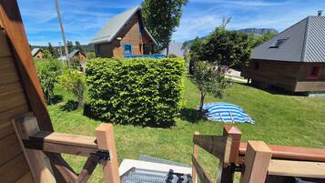 Une terrasse en bois avec un barbecue en métal, une pelouse, une maison en bois avec une porte bleue et un arbre aux fleurs roses.