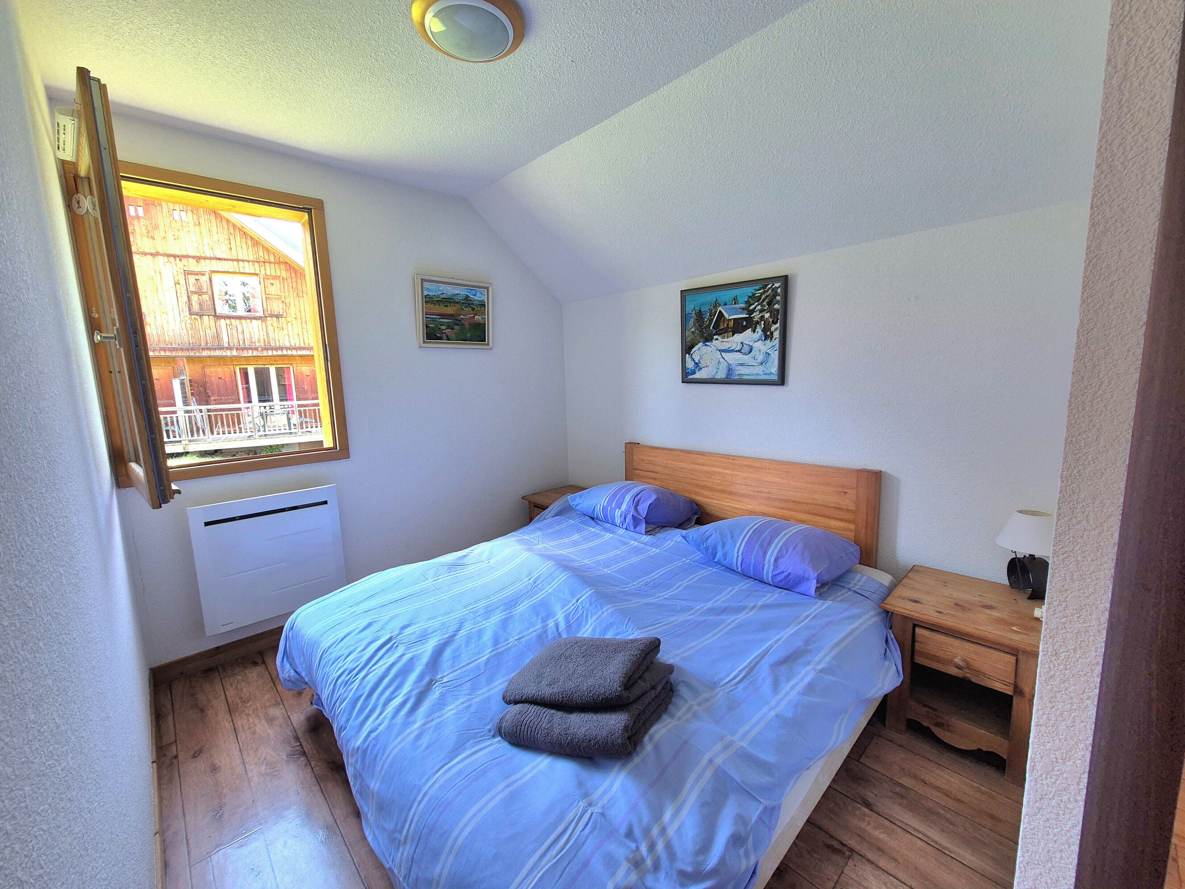 Ein Schlafzimmer mit einem Bett, einem Nachttisch, einem Fenster mit Blick auf ein Holzhaus und zwei an der Wand befestigten Bildern.