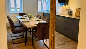 Comedor con una mesa de madera dispuesta para cuatro personas, rodeada de sillas. Se observan dos ventanas con marcos blancos, una zona de cocina con campana extractora negra y un pilar de madera en primer plano.