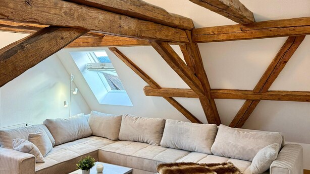 Habitación tipo loft con vigas de madera a la vista, sofá modular, mesa de centro y ventana pequeña con vistas.