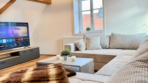 Un dormitorio tipo loft acogedor con vigas de madera a la vista, un ventanal grande, un conjunto de sofás, un televisor de pantalla plana y una pequeña planta.